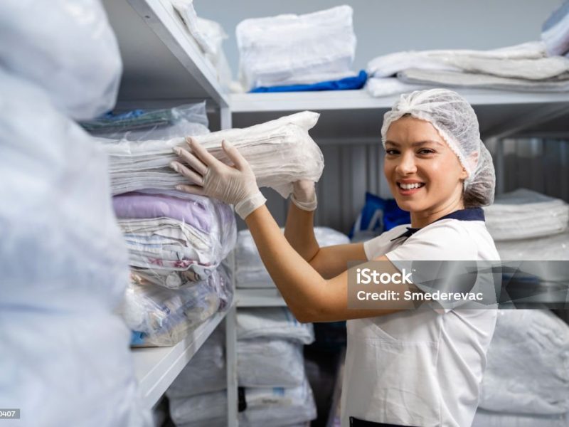 Female laundry worker stacking shelves with clean clothes and linen in storage room.