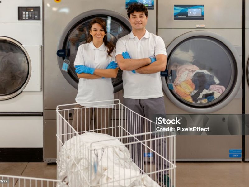 Two young entrepreneurs posing for the camera in their laundry service shop, surrounded by clean clothes and laundry machines, representing their hard work and dedication.